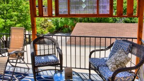 A covered outdoor seating area with wicker chairs and a glass table, overlooking a brick building and lush greenery. - Bard's Inn (Cedar City)