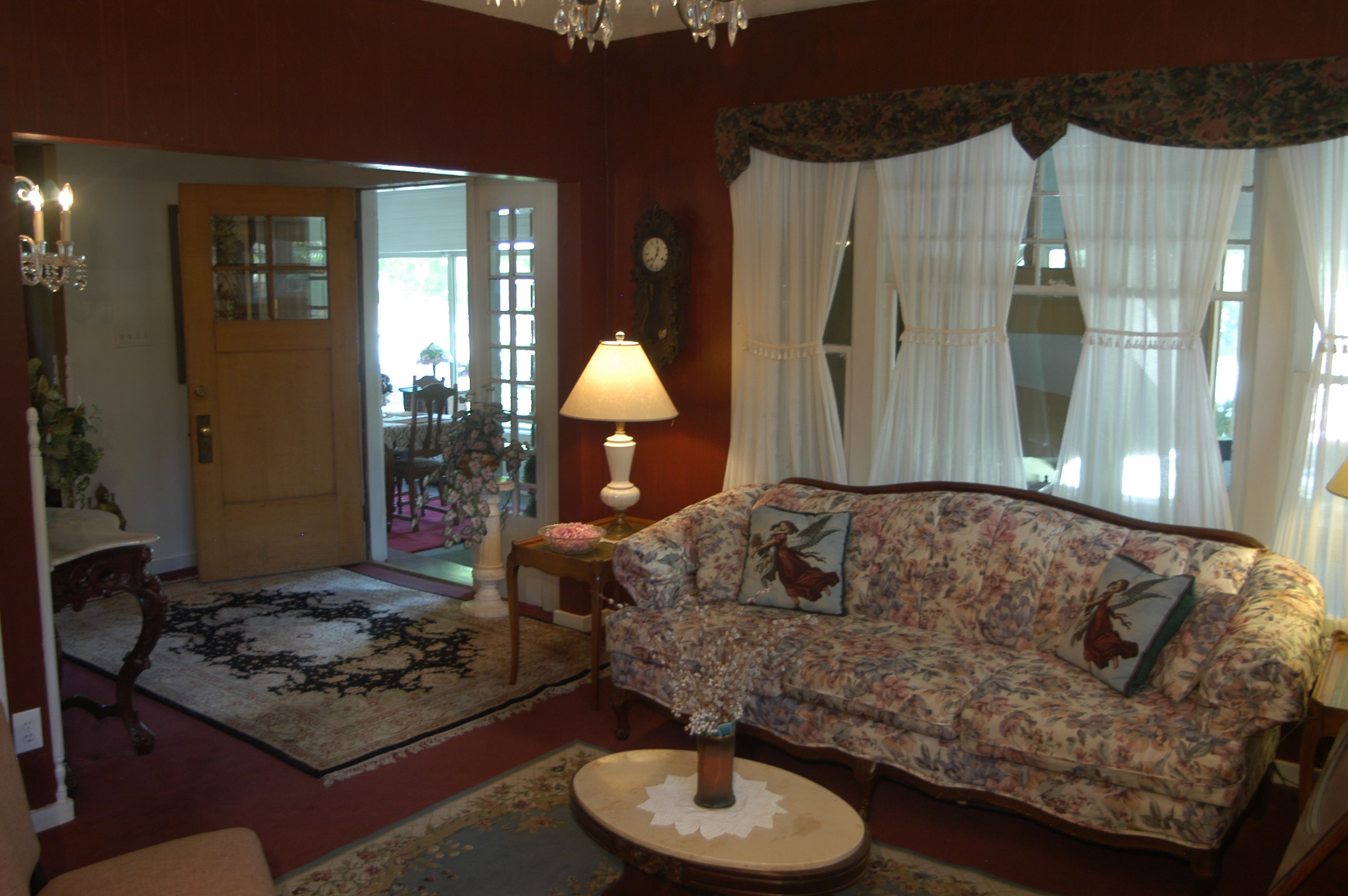 A living room with a floral sofa, a round coffee table, a lamp, and a large window with sheer curtains.