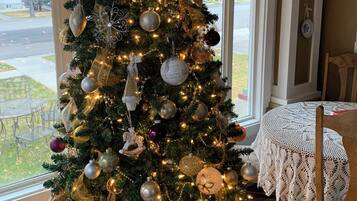 Un sapin de Noël décoré de guirlandes et de lumières, placé dans une pièce dotée d’une grande fenêtre, d’une chaise en bois et d’une nappe en dentelle.