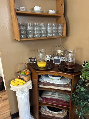 A beverage station with a fruit basket, a tray of bananas, and two pitchers labeled "Orange Juice" and "Apple Juice" on a wooden table.