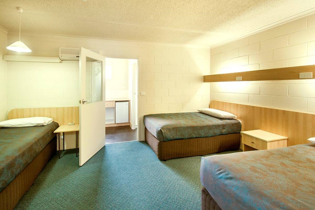 A hotel room with two single beds, a small desk, a chair, a bedside table, and a wall-mounted air conditioning unit.