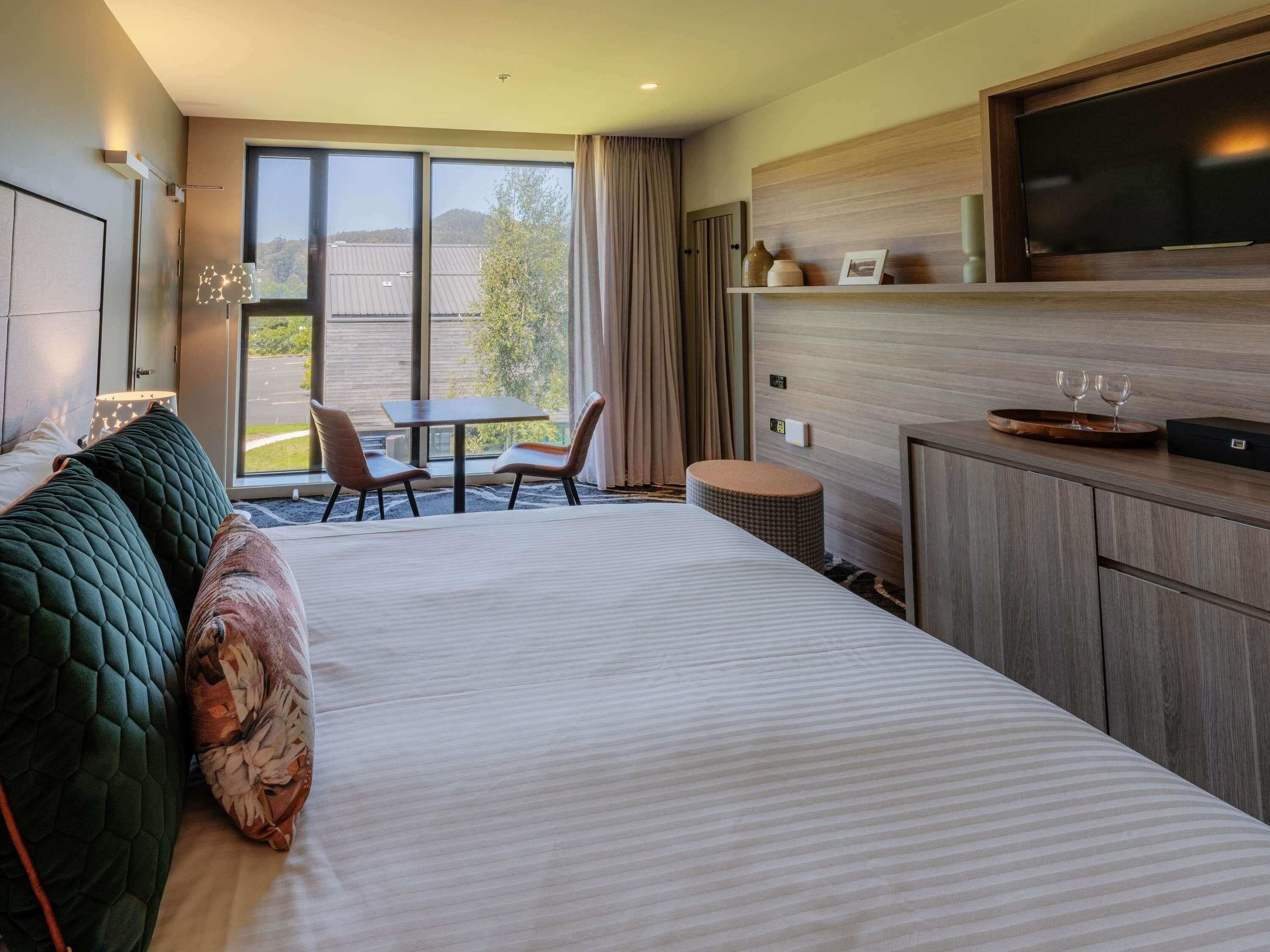 A hotel room with a large bed, a dining table with chairs, a television, and a view of the outdoors.