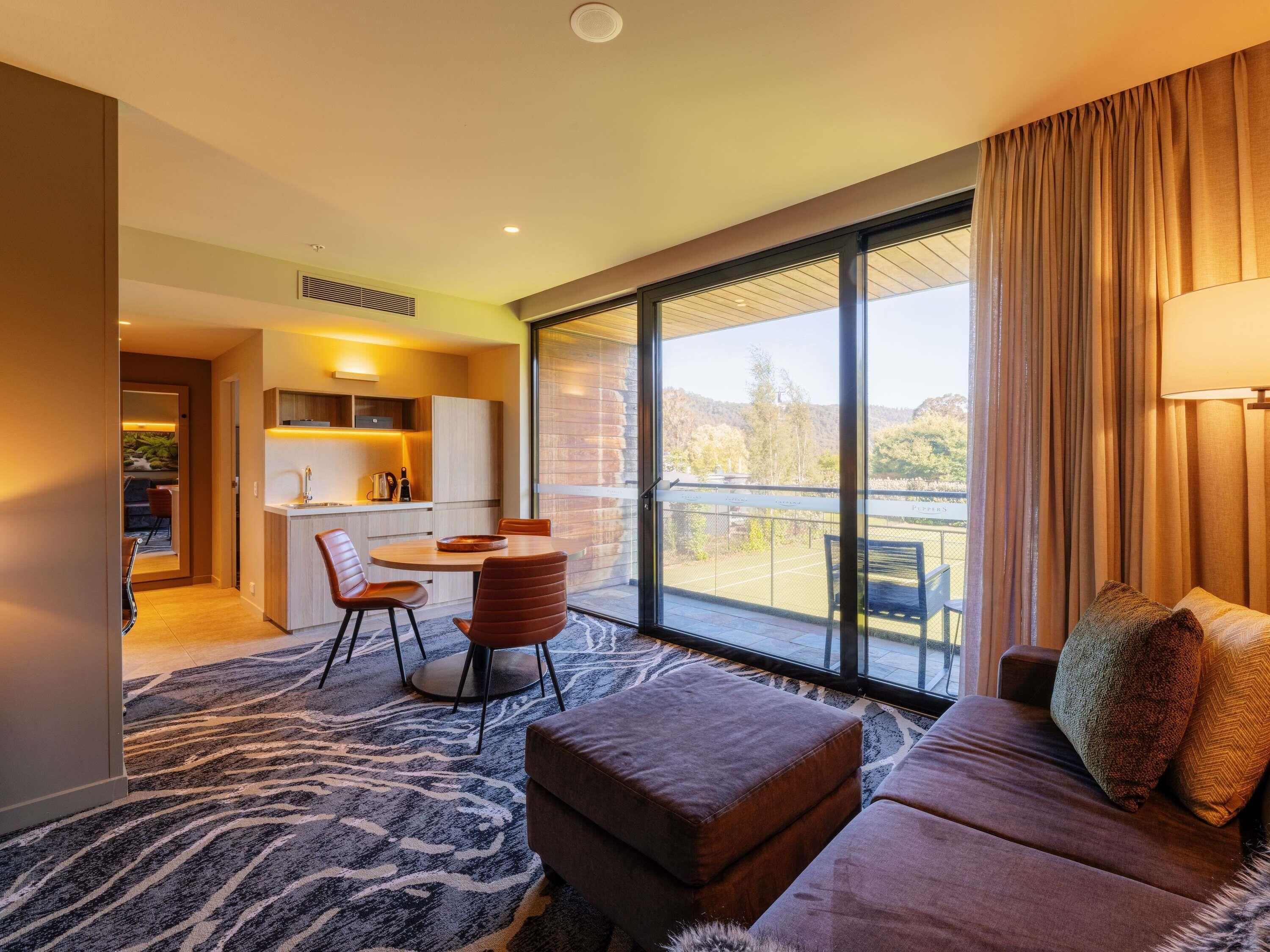 A modern hotel room with a balcony, a sofa, a dining area, and a kitchen.