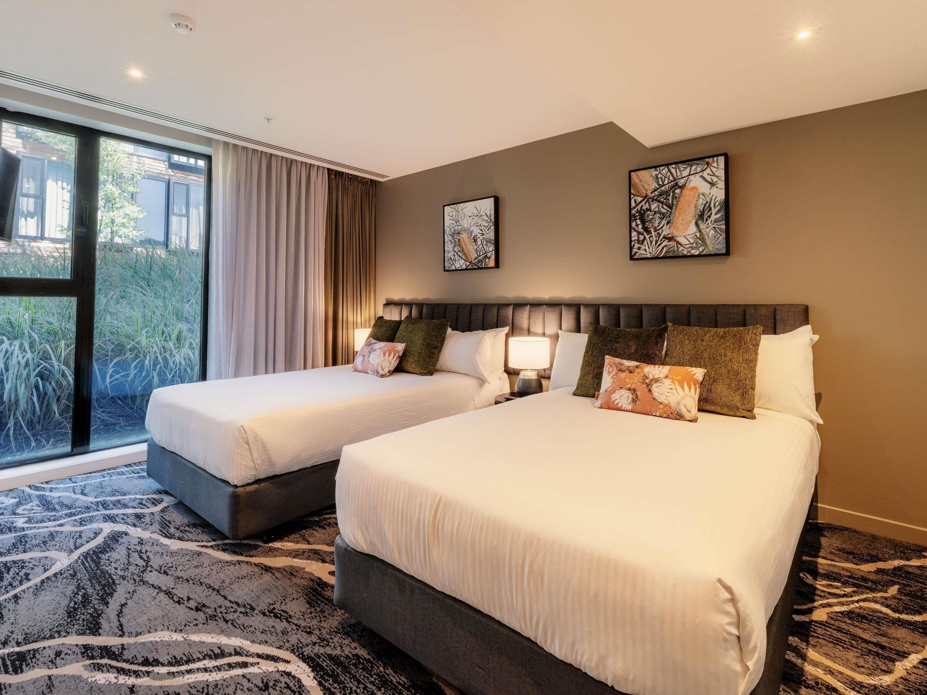 A hotel room with two beds, a large window with curtains, and two wall-mounted artworks.