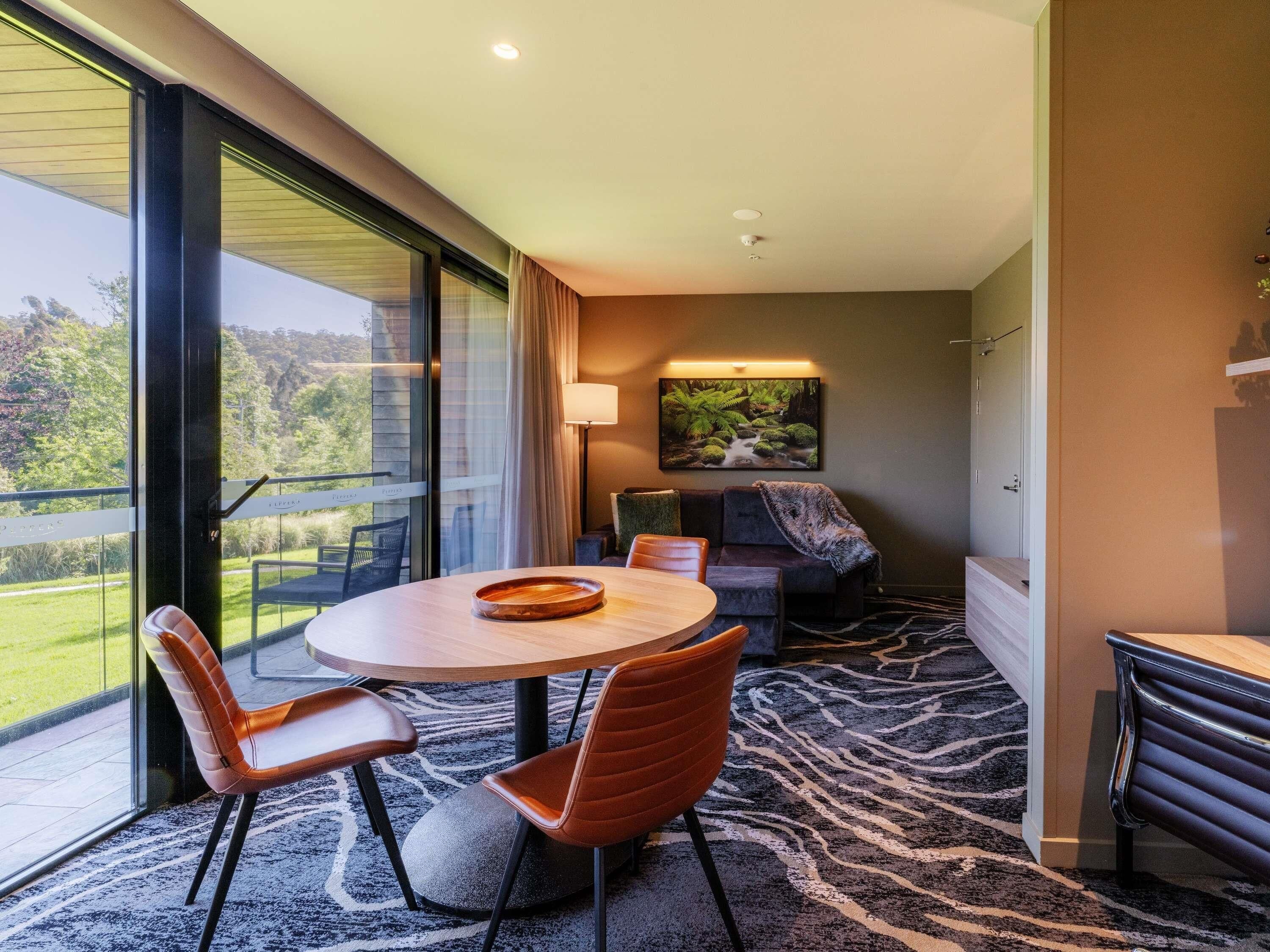 A modern hotel room with a large window, a round dining table, two chairs, a sofa, and a painting of a forest stream.