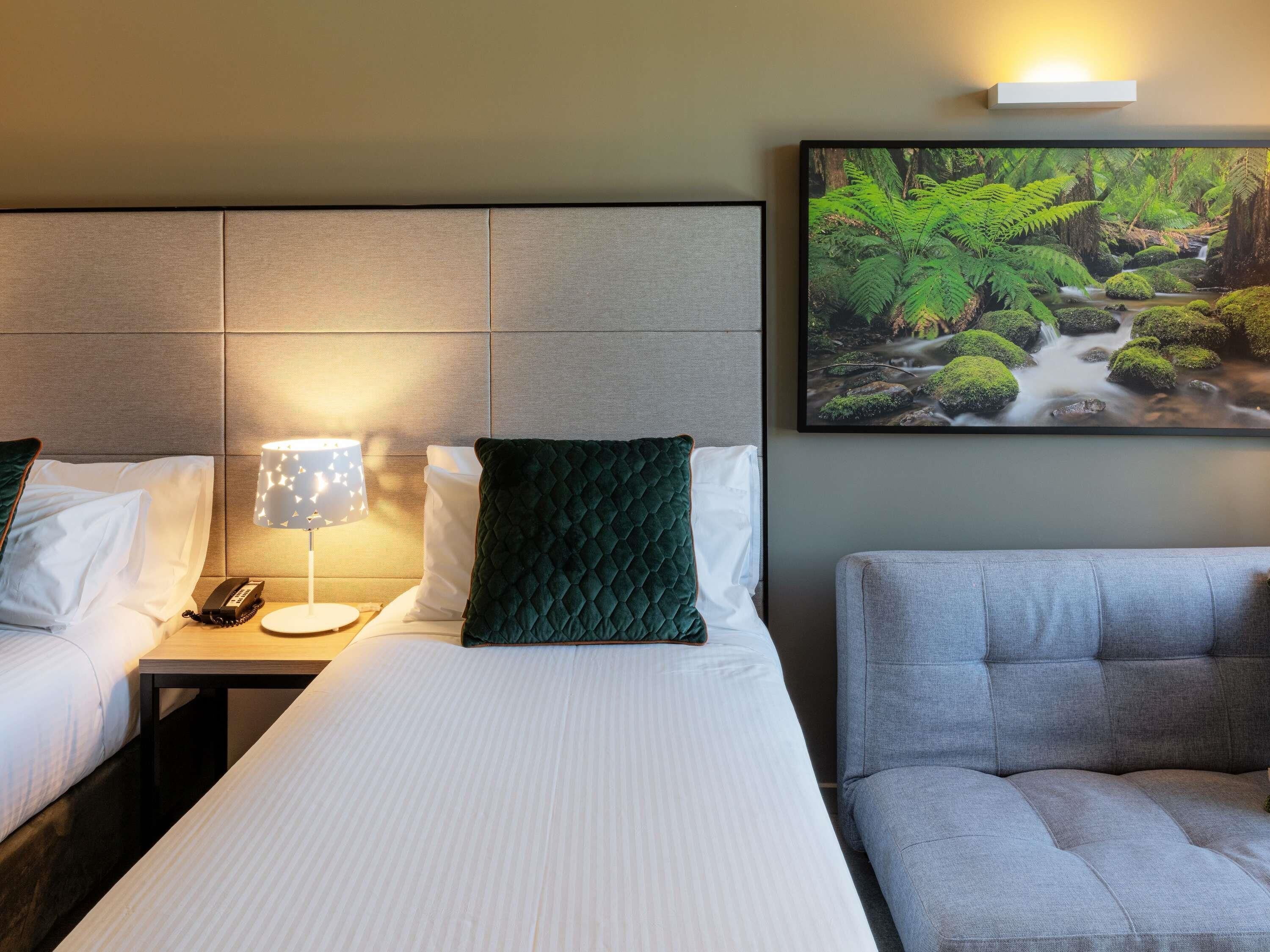 A hotel room with a bed, bedside table, lamp, phone, and a framed picture of a forest stream.