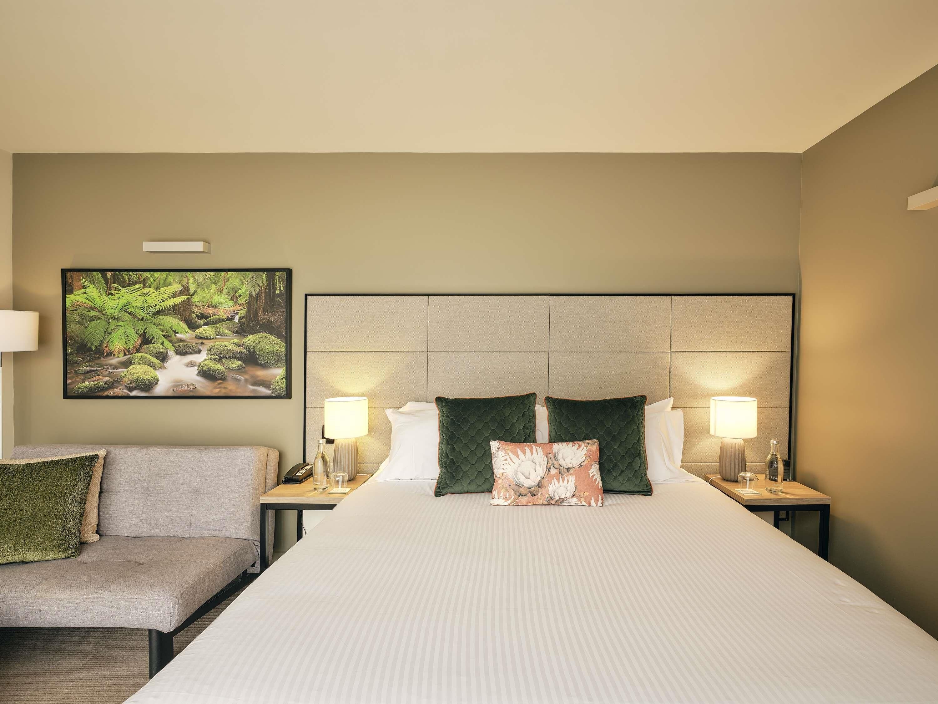 A hotel room with a large bed, two bedside tables, a nightstand, a sofa, and a wall-mounted TV.