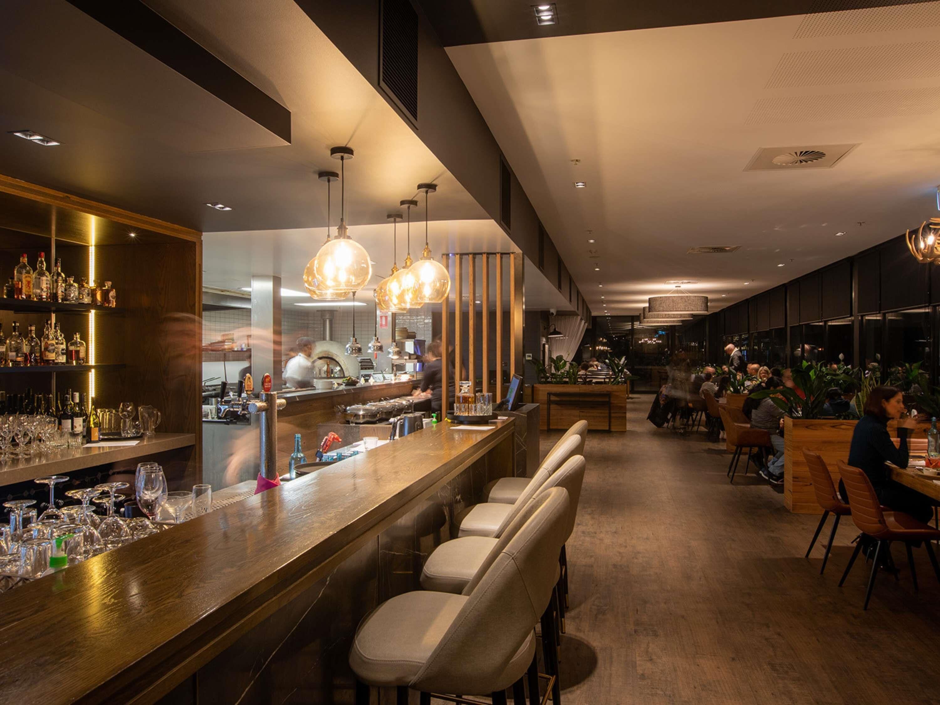 A bar area with stools, a counter with bottles, and a kitchen in the background.