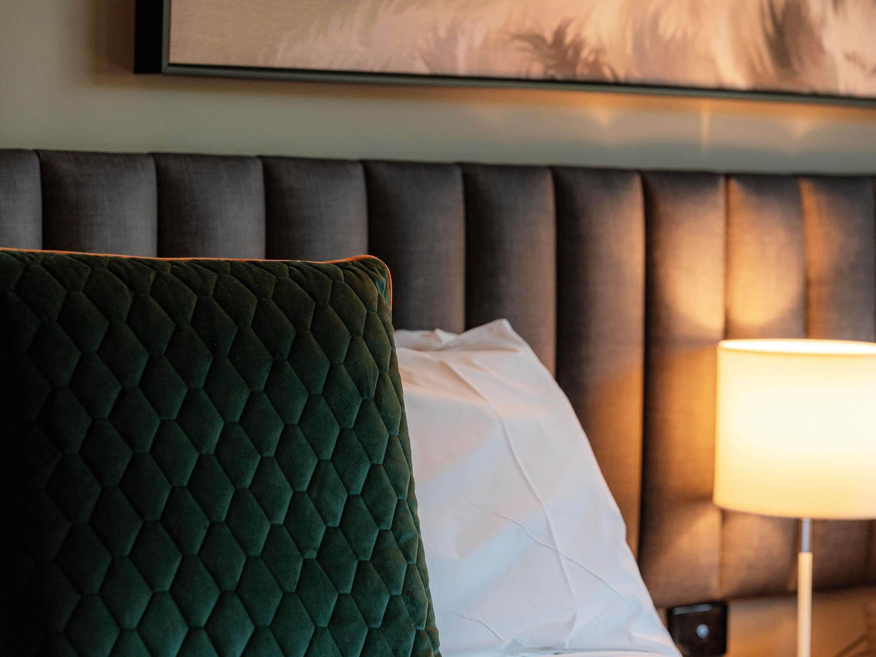 Habitación de hotel con cabecera oscura, una butaca tapizada de verde, almohada blanca y lámpara de noche.