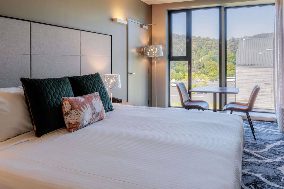 Habitación de hotel con una cama grande, dos sillas, una mesa, una lámpara y vistas al exterior.