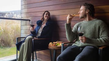 Two people seated on a bench outside a building with a wooden exterior, holding glasses of red wine.