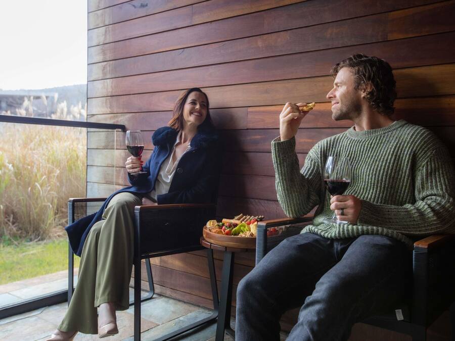 Two people seated on a bench outside a building with a wooden exterior, holding glasses of red wine.