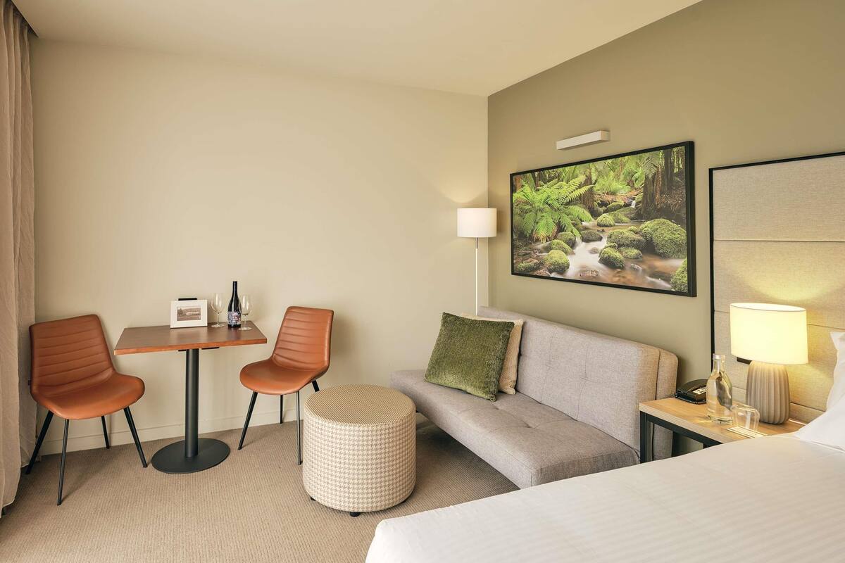 Una habitación de hotel con una cama, un sofá, dos sillones, una mesita con una botella y vasos, una lámpara, un cuadro en la pared y un reposapiés a cuadros.