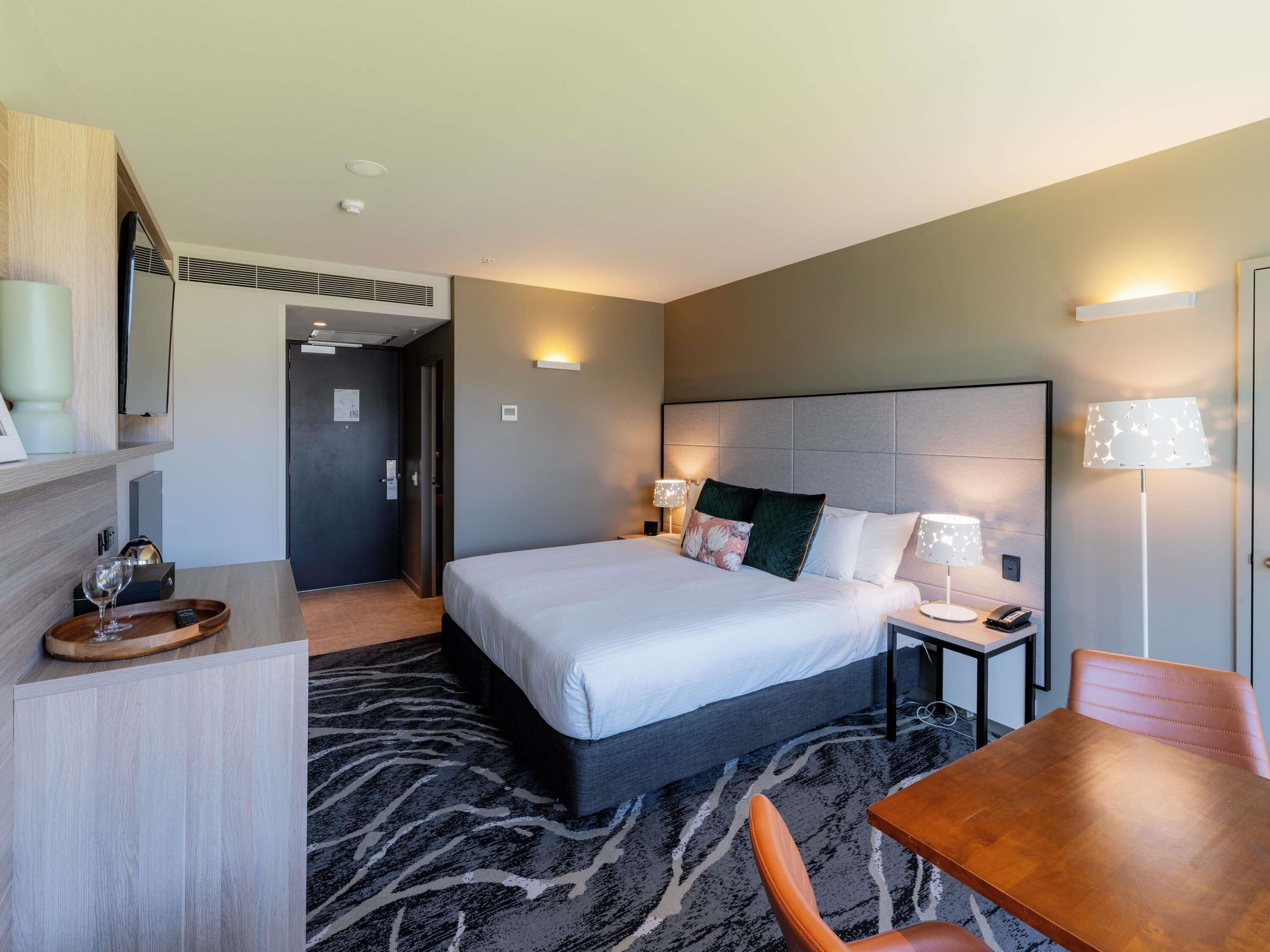 A hotel room with a bed, bedside tables, a desk, a chair, and a carpeted floor.