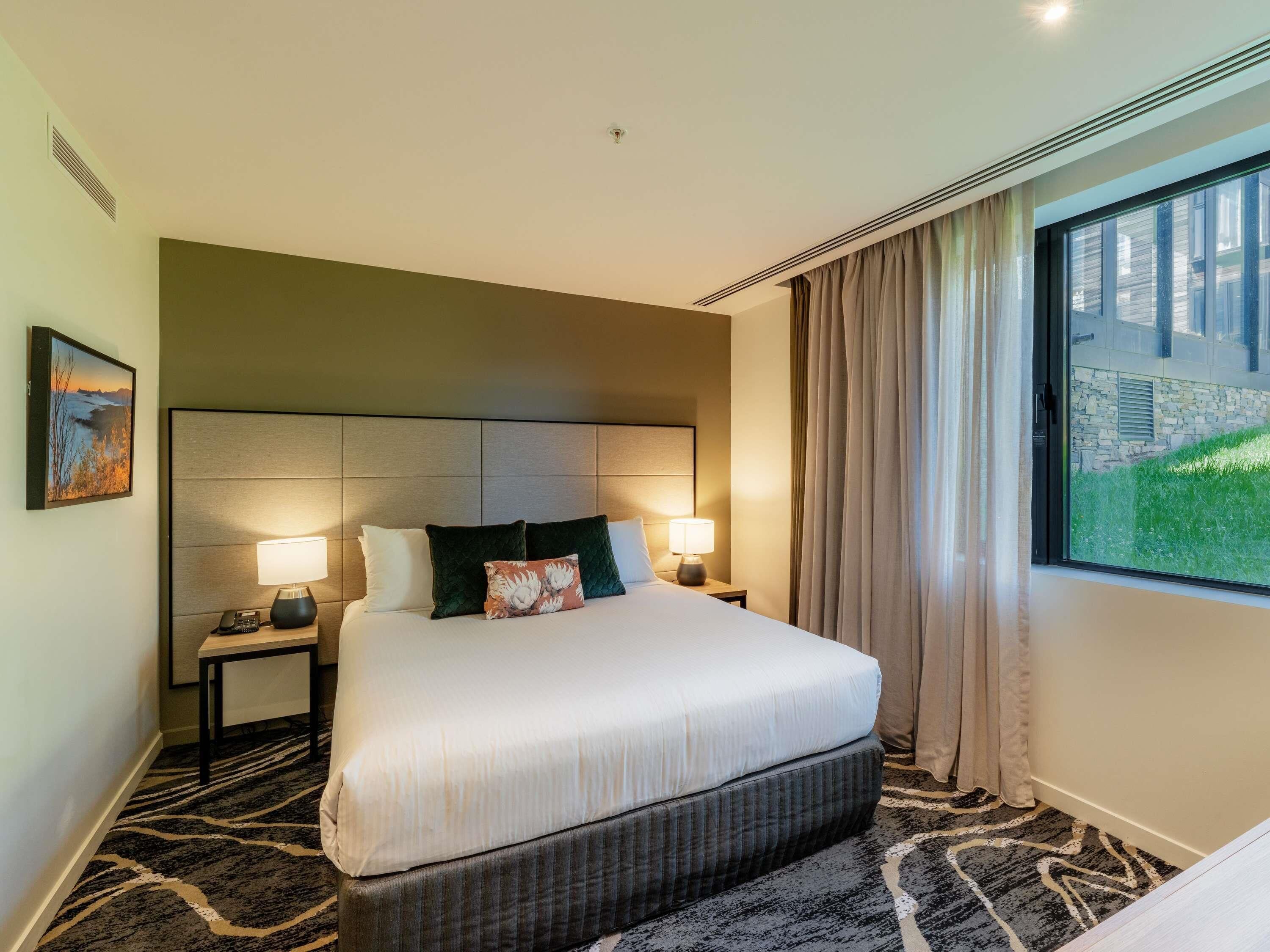 Habitación de hotel con una cama grande, dos mesitas de noche con lámparas, un cabecero, una ventana con cortinas y un cuadro en la pared.
