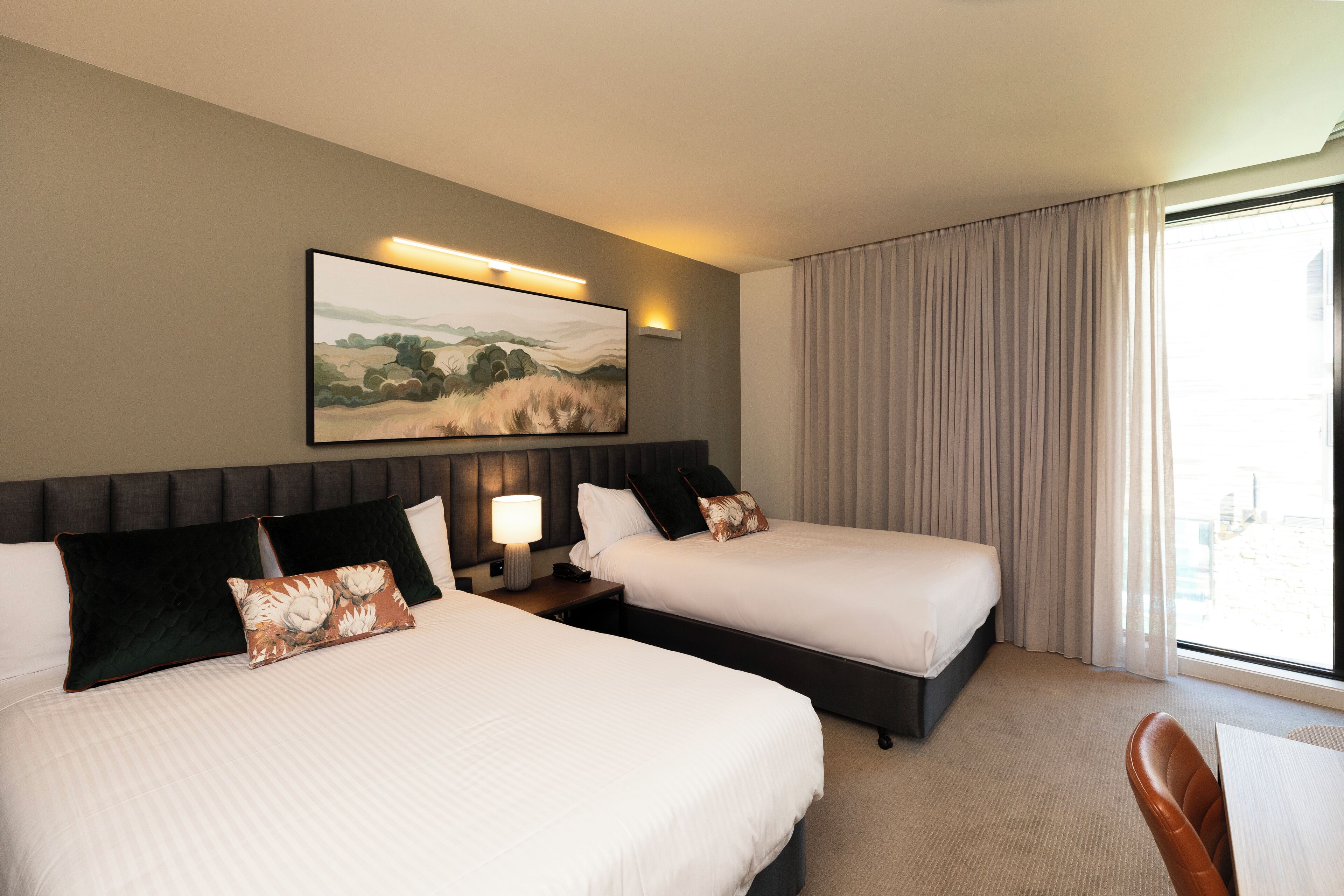 Habitación de hotel con dos camas, un cuadro de paisaje grande, una ventana con cortinas y vistas a la ciudad.
