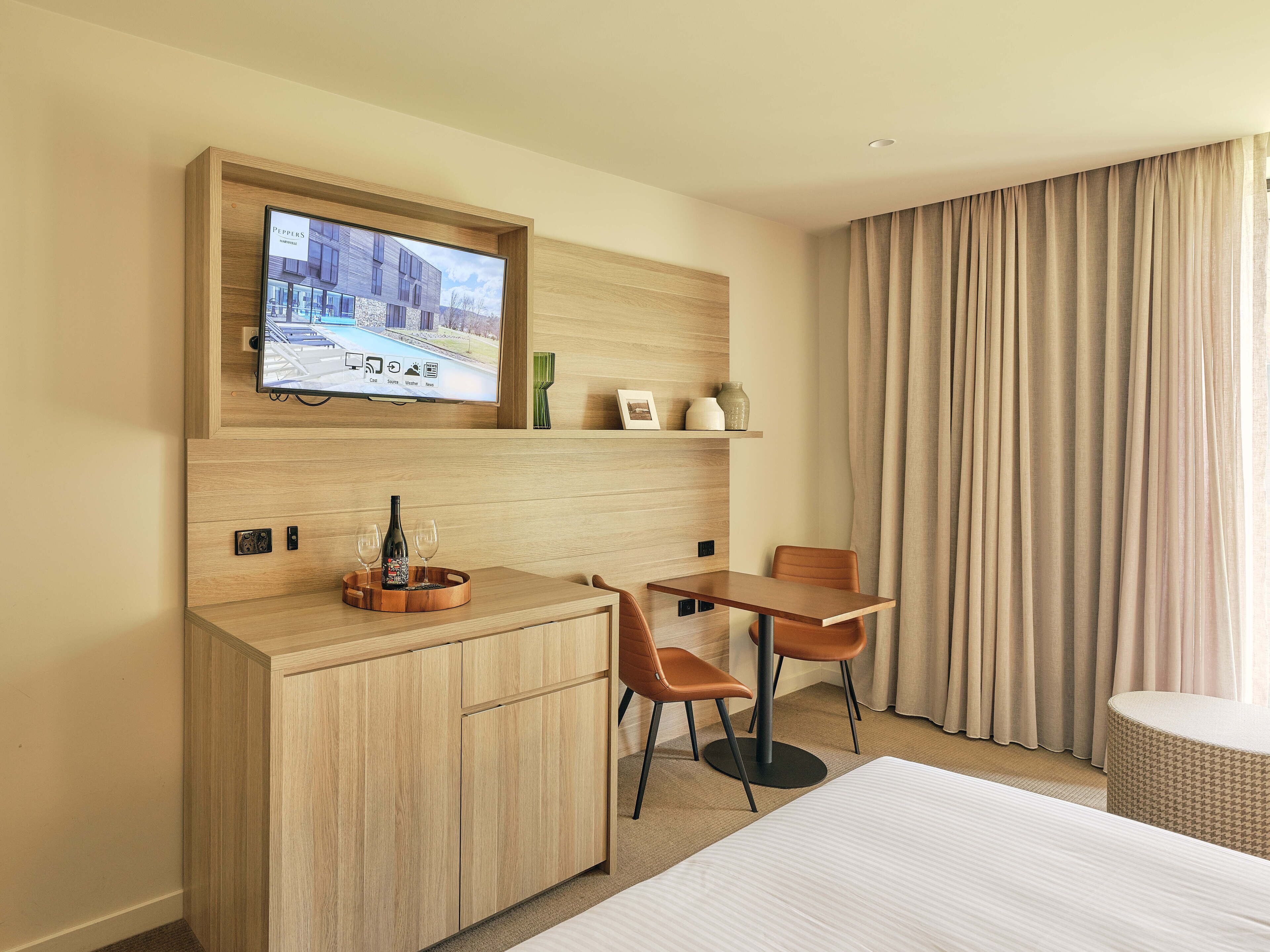 Ein Hotelzimmer mit einem Holztisch, zwei Stühlen, einem Fernseher über dem Tisch, einem Bett und einem großen Fenster mit Vorhängen.