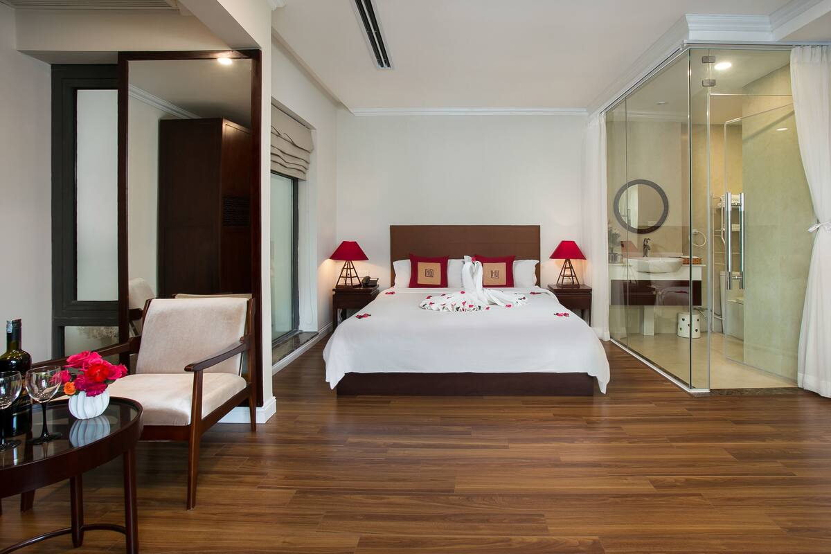 A hotel room with a large bed, a sitting area with a chair, a glass-enclosed shower, and a dining table with a vase of flowers.