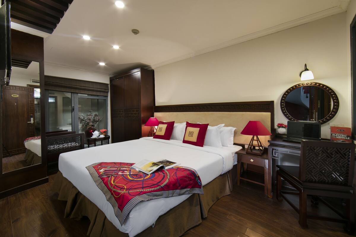 A hotel room with a large bed, a bedside table with a lamp, a mirror, and a desk with a chair.