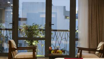 A hotel room with two wooden chairs, a bed with a red and yellow bedspread, and a balcony with a table holding a fruit plate and a potted plant.