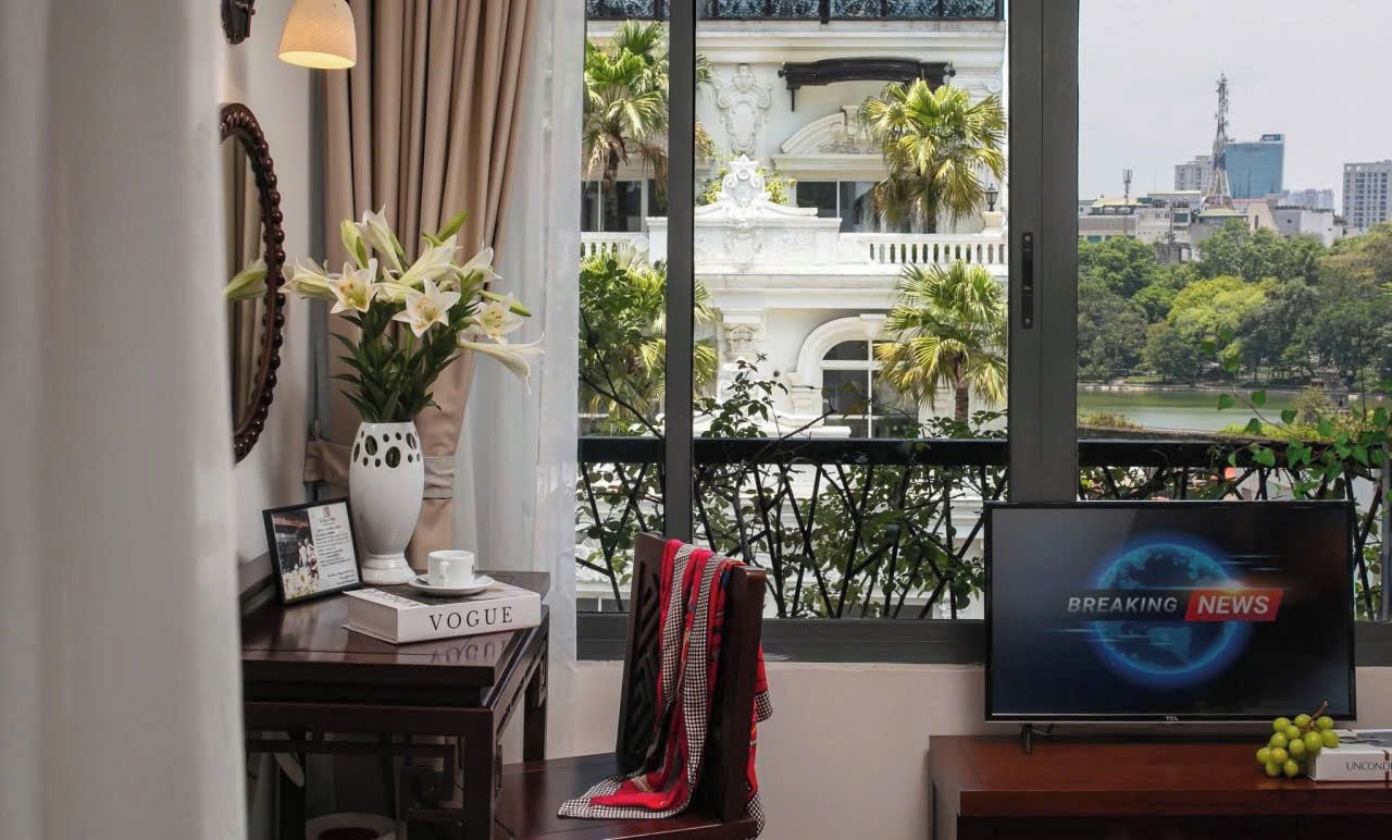 Ein Zimmer mit einem großen Fenster, einem Fernseher, der 'BREAKING NEWS' zeigt, einer Vase mit Blumen, einem gerahmten Bild und einem Buch mit dem Titel 'VOGUE'.