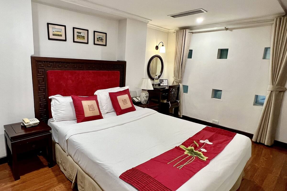 A hotel room with a large bed, a wooden headboard, a nightstand with a phone, a desk with a lamp, and a mirror.
