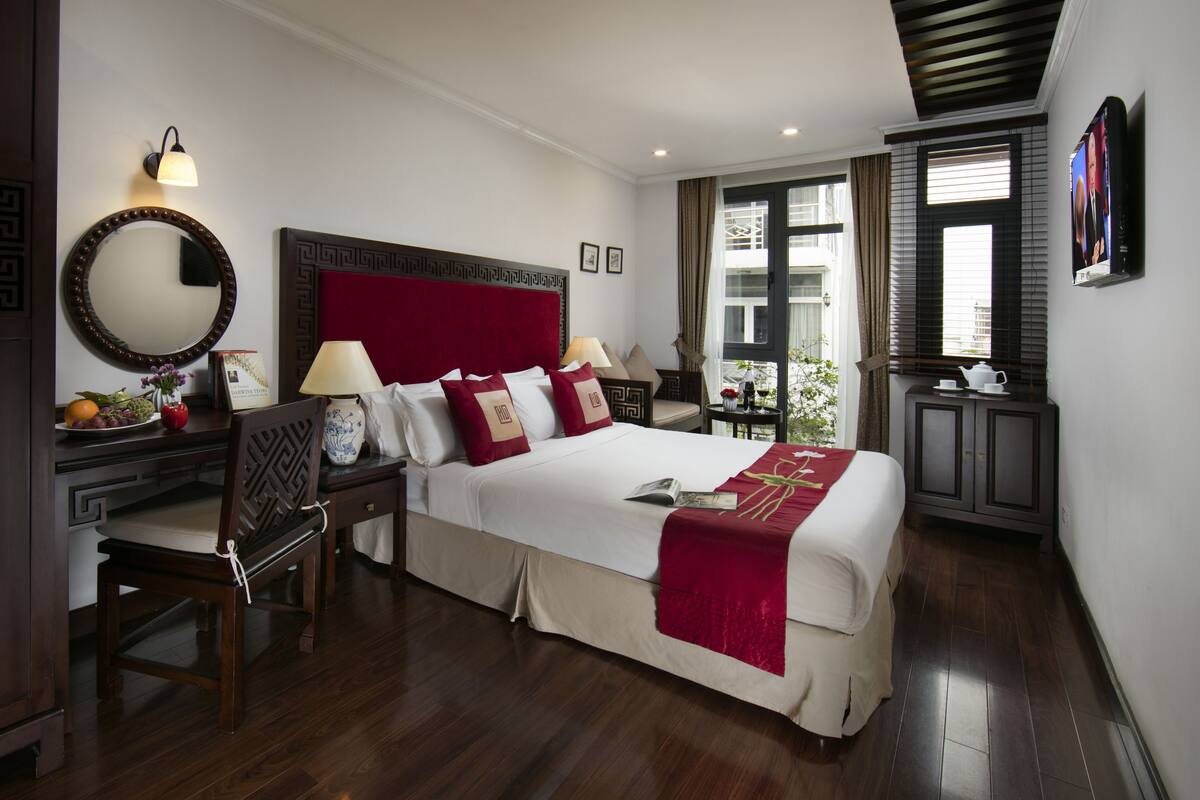 A hotel room with a large bed, bedside tables, a desk, and a chair. There is a window with curtains and a view of the outside.