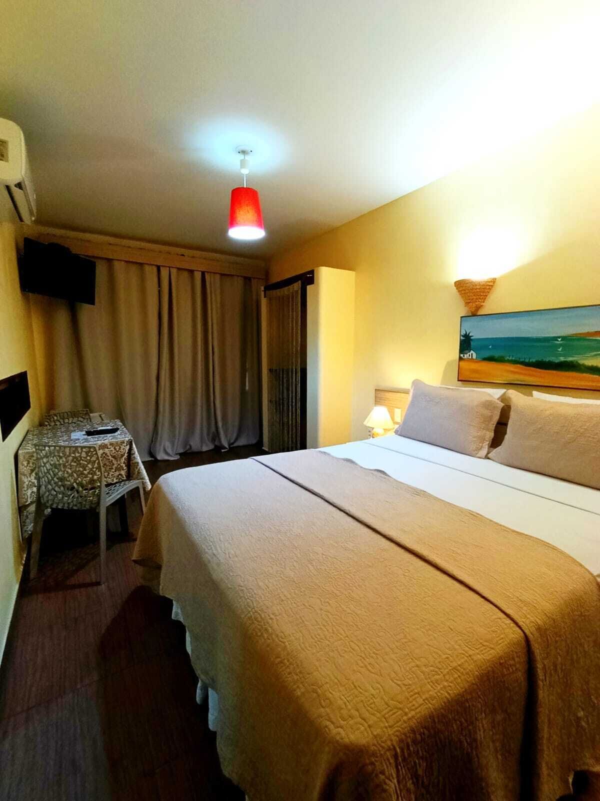 A hotel room with a bed, a desk with a chair, a TV, a painting on the wall, and a ceiling light.