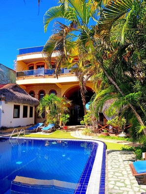 A poolside view of a hotel with a clear blue pool, a thatched-roof structure, and lush tropical vegetation. - Pousada Presidente Hotel (Aracati)