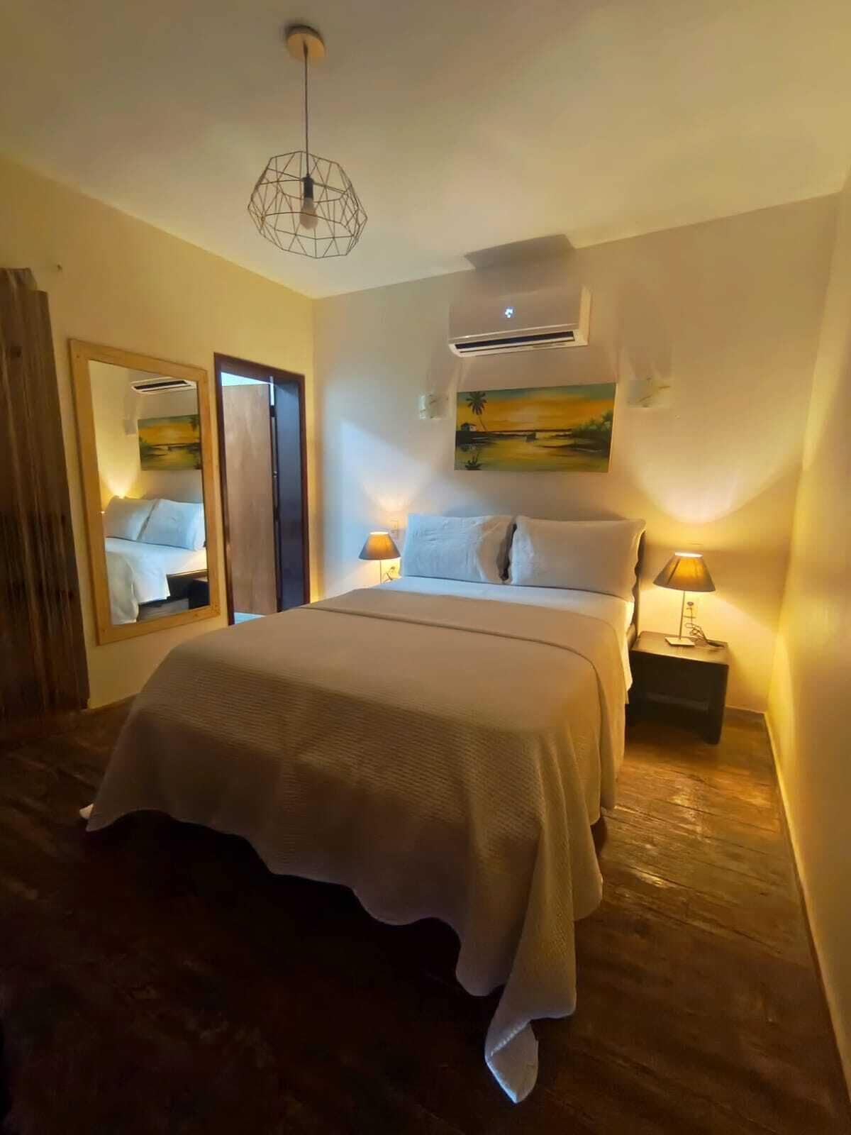 Un dormitorio con una cama, dos mesitas de noche, aire acondicionado de pared, un cuadro y un espejo.