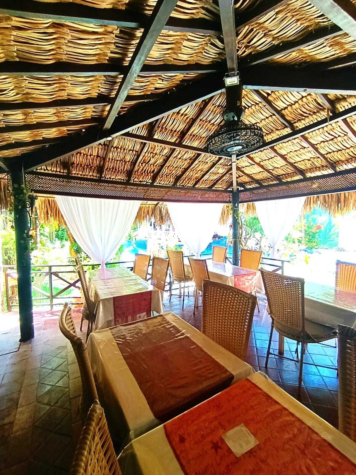 Un área de comedor al aire libre con mesas y sillas, techo de paja y vista a la vegetación.