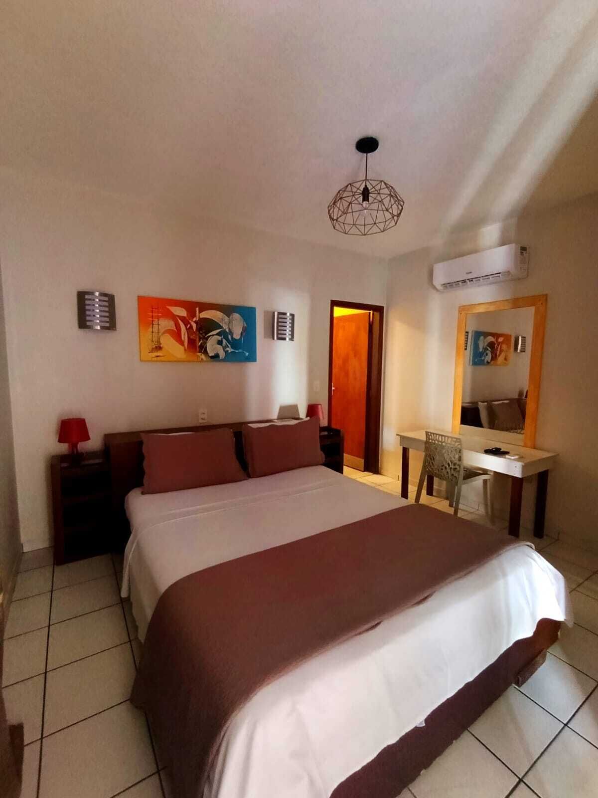 Quarto de hotel com cama, escrivaninha, cadeira, abajur vermelho, pintura e ar condicionado, além de uma porta.