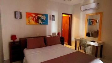 Quarto de hotel com cama, escrivaninha, cadeira, abajur vermelho, pintura e ar condicionado, além de uma porta.