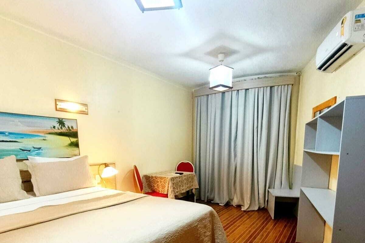 A hotel room with a bed, a desk, a chair, a table, a wardrobe, and a painting on the wall.