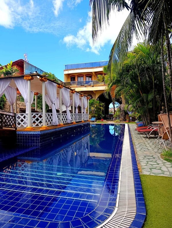 A swimming pool with a tiled edge, a covered seating area with white curtains, and a building with balconies in the background. - Pousada Presidente Hotel (Aracati)