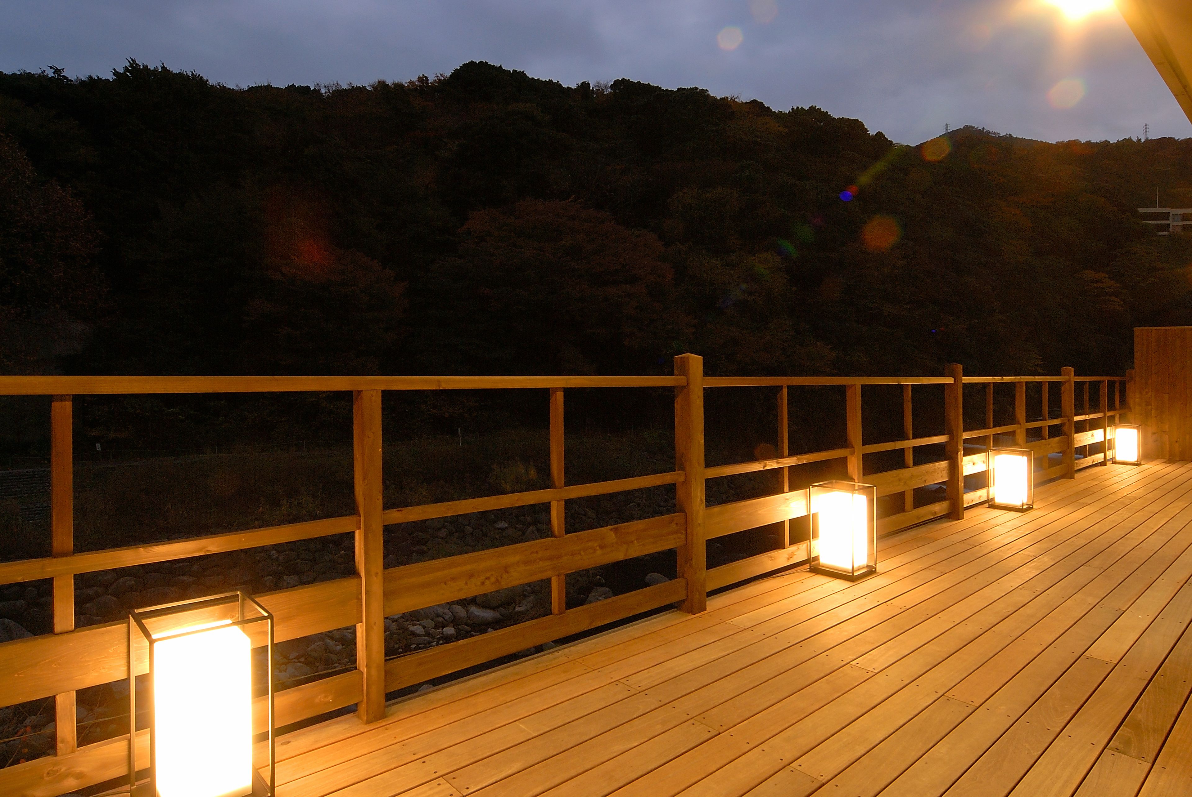Een houten terras met balustrades en verlichte lantaarns, met uitzicht op een bebost helling.
