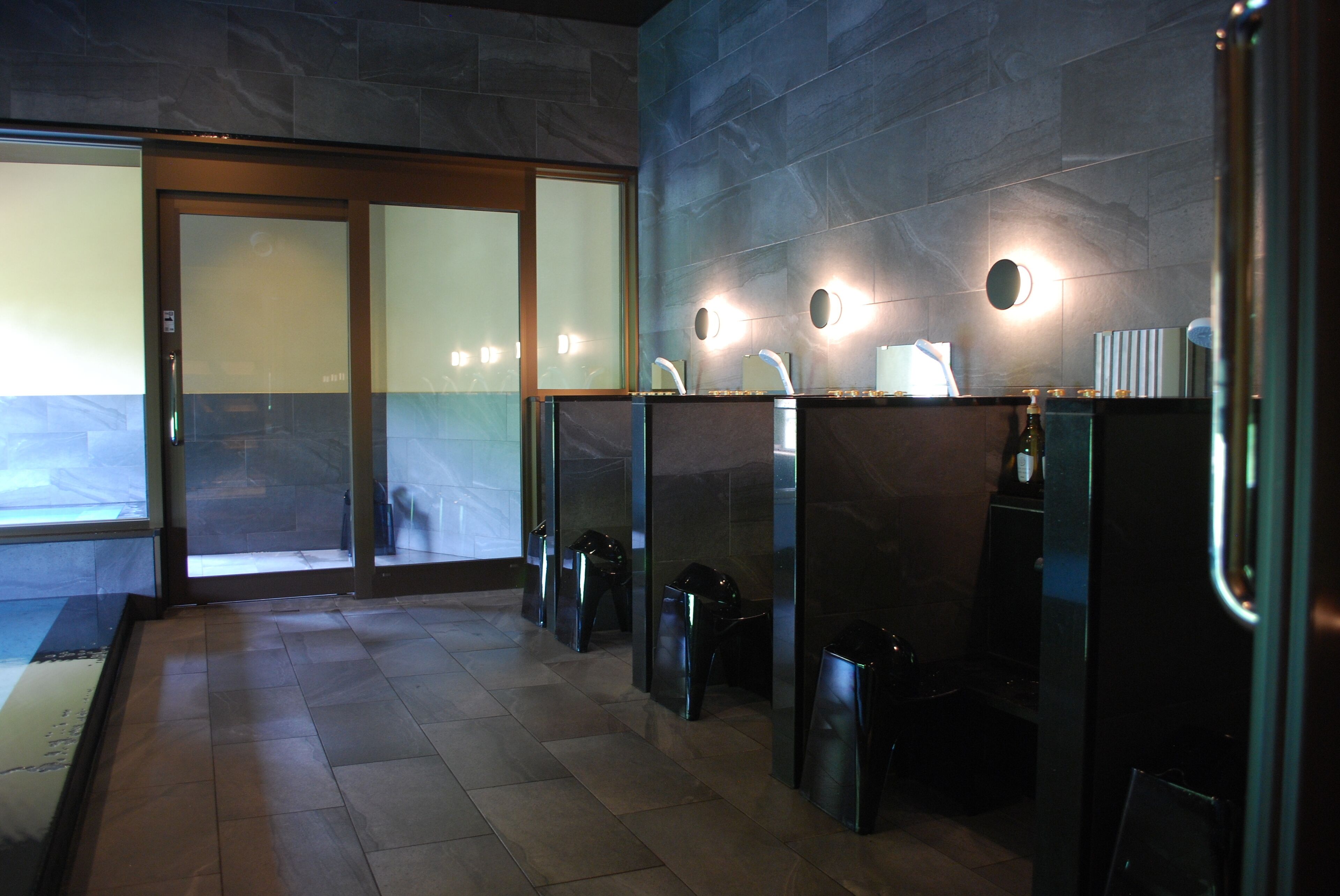 A modern restroom with glass partitions, a tiled floor, and wall-mounted lights.