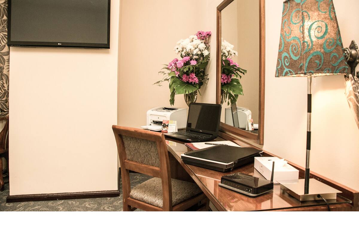 A hotel room with a desk, laptop, chair, mirror, and a lamp.