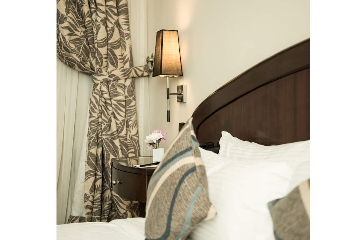 A hotel room with a bed, pillows, a nightstand, a lamp, and curtains.