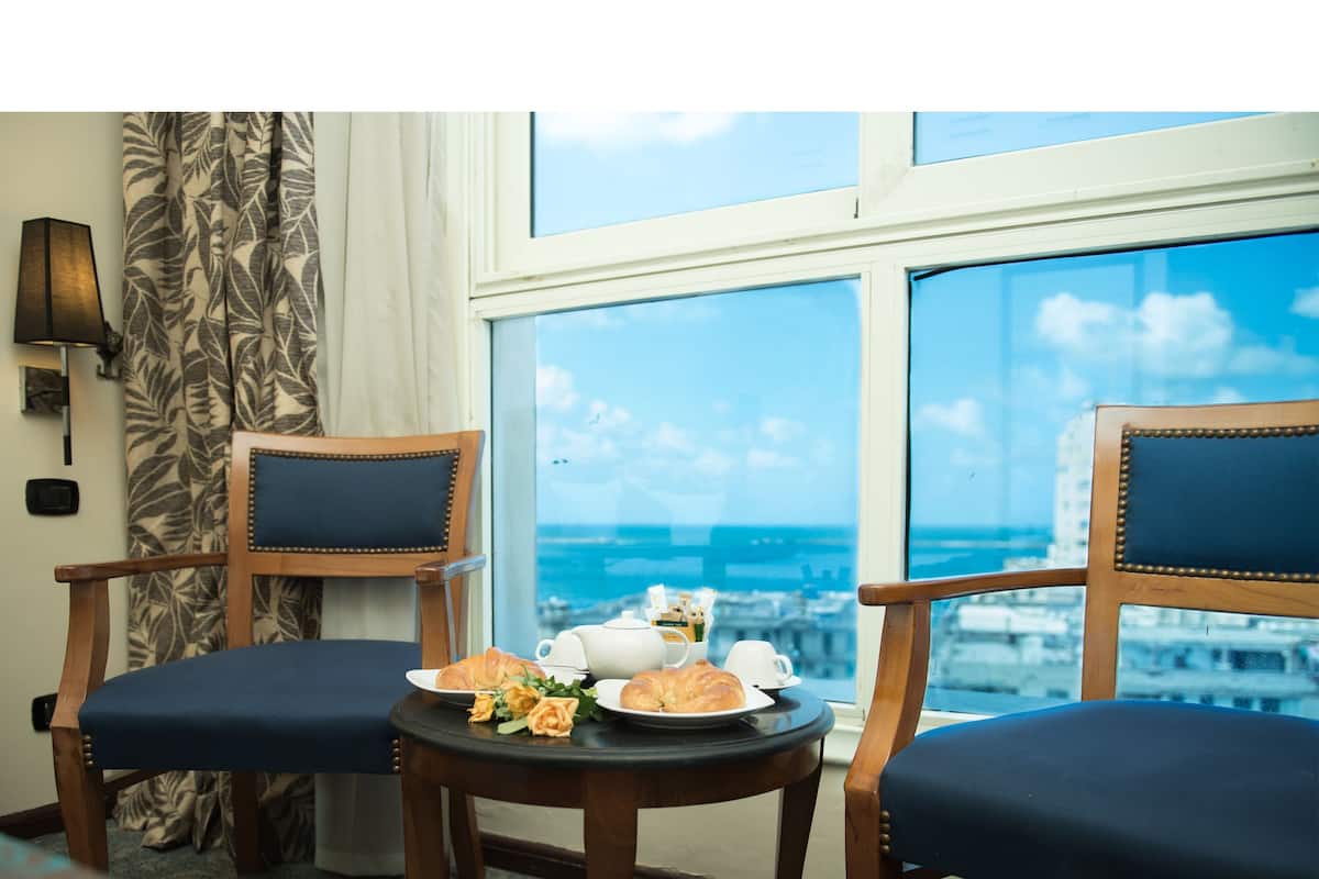 A hotel room with two wooden chairs, a small table with plates of food, a teapot, and a cup. A window offers a view of the cityscape.