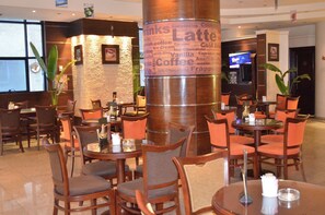 A café with round tables and chairs, a large pillar with drink names, and a TV screen in the background. - Cherry Maryski Hotel (Alexandria)