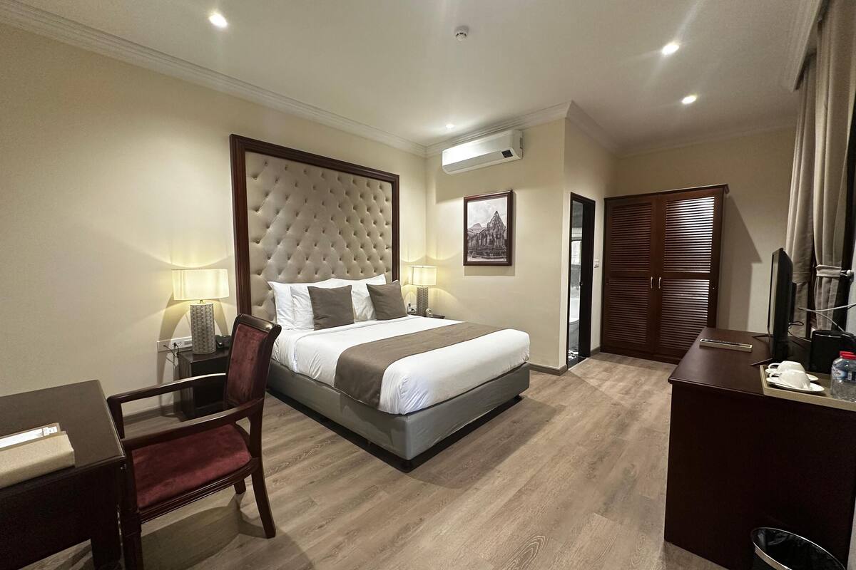 A hotel room with a large bed, a desk, a chair, a TV, and an air conditioning unit.