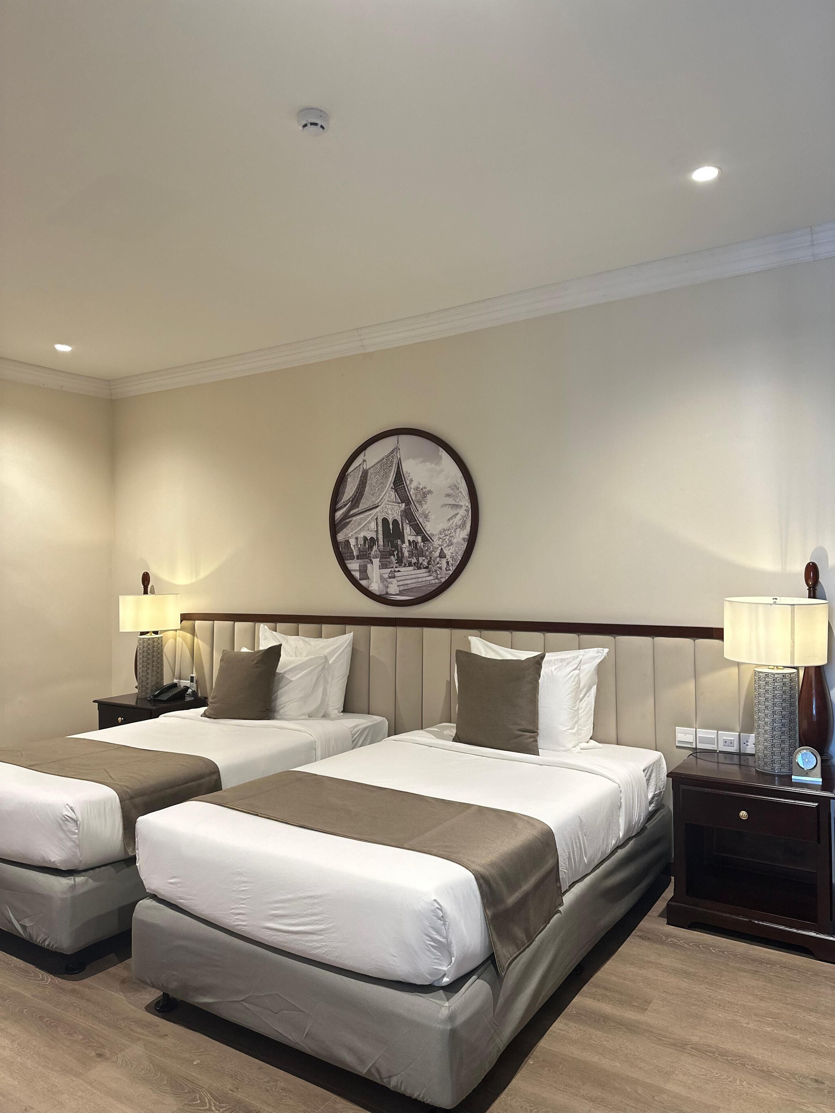 A hotel room with two beds, a nightstand with a lamp, a wall-mounted picture, and a ceiling light.