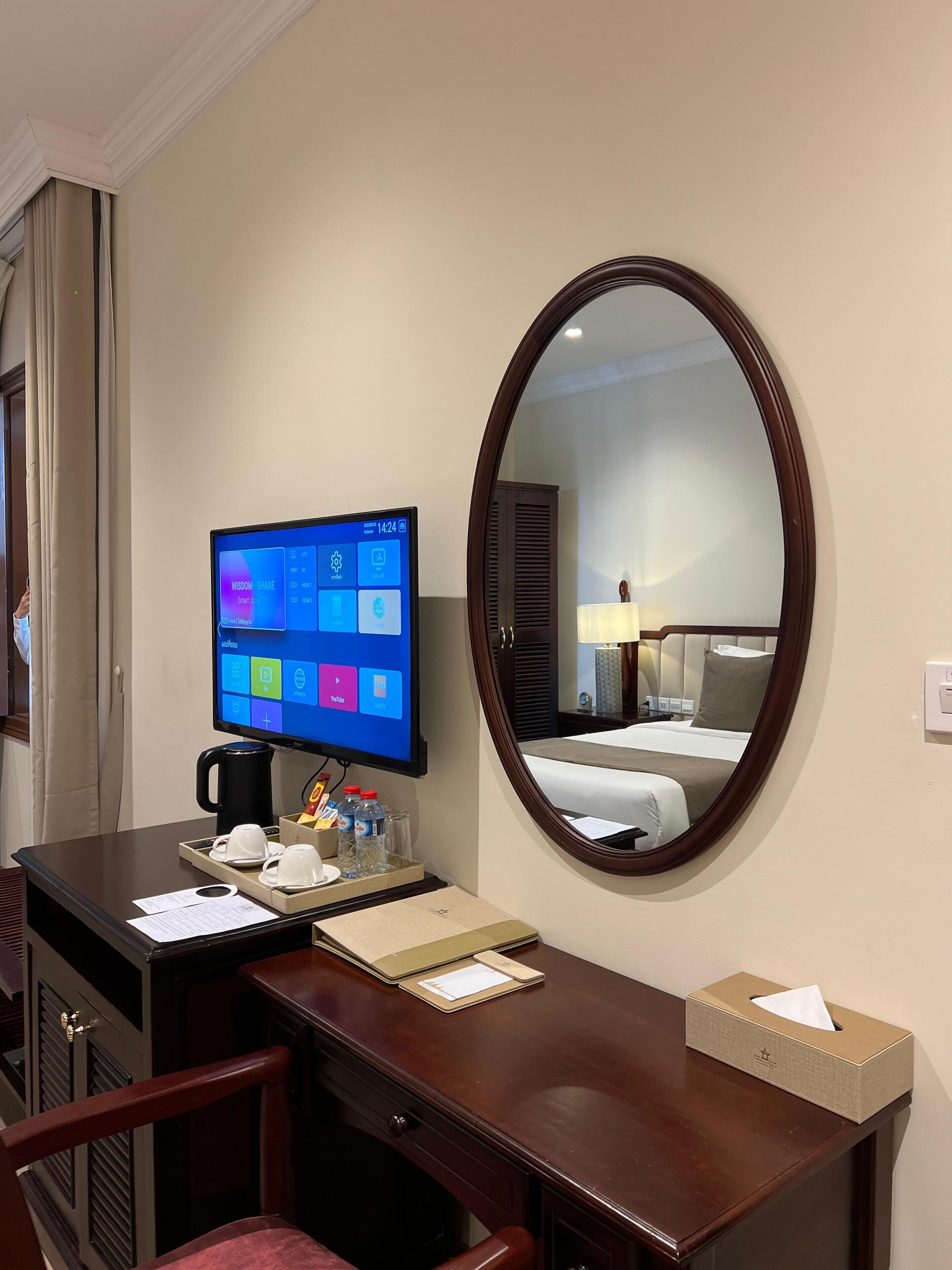 Habitación de hotel con escritorio, monitor de computadora, espejo, cama y mesita de noche con lámpara y caja de pañuelos.