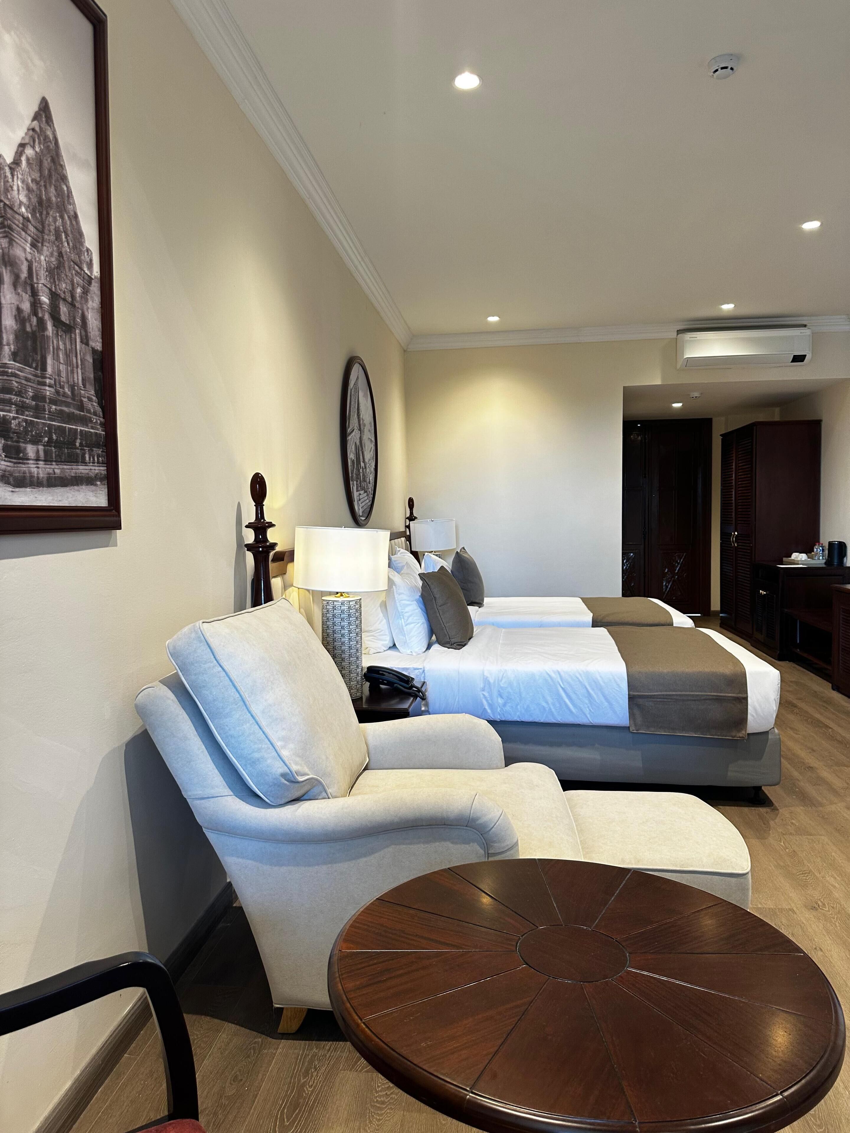 Une chambre d’hôtel avec un canapé, deux lits, une table basse ronde, une lampe et un tableau encadré au mur.