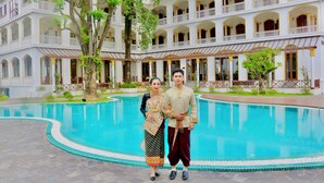 A couple standing by a pool with a multi-story building in the background. - Champa Palace Hotel (Pakse)