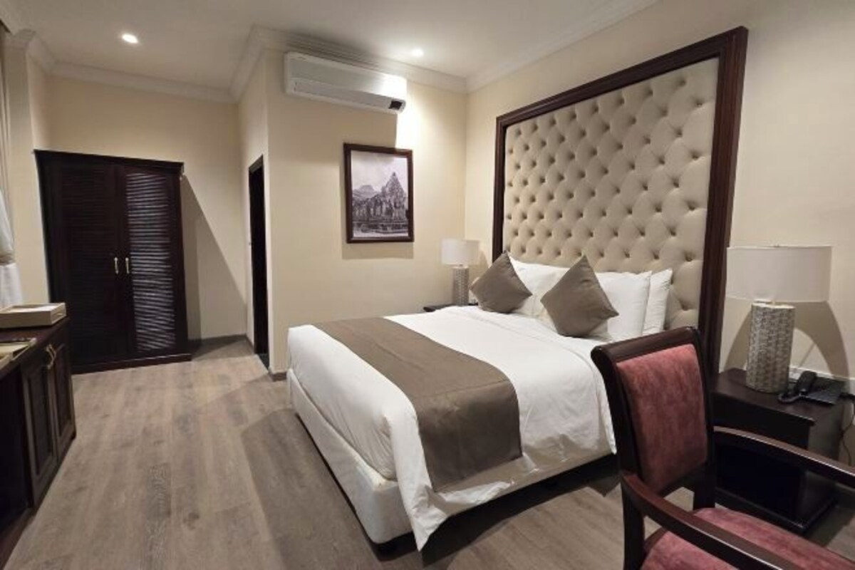 A hotel room with a large bed, a wooden wardrobe, a bedside table with a lamp, and a framed picture on the wall.