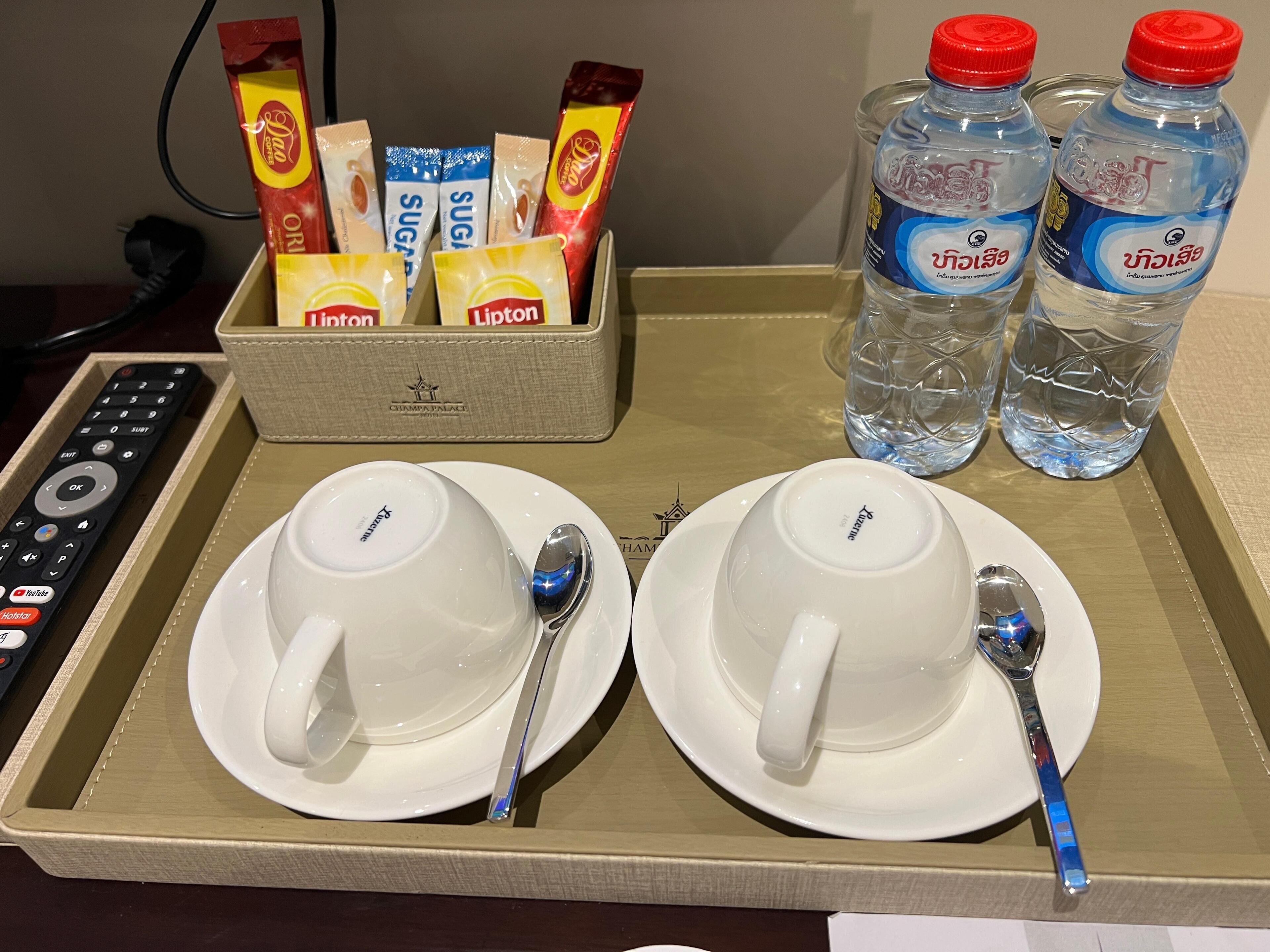 A tray with two white cups, two spoons, two bottles of water, a remote control, and a box of Lipton tea packets.