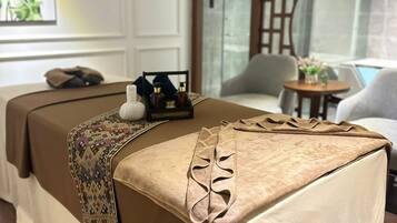 Une table de massage recouverte d’une couverture à motifs, une petite table avec des bouteilles et un élément décoratif mural.