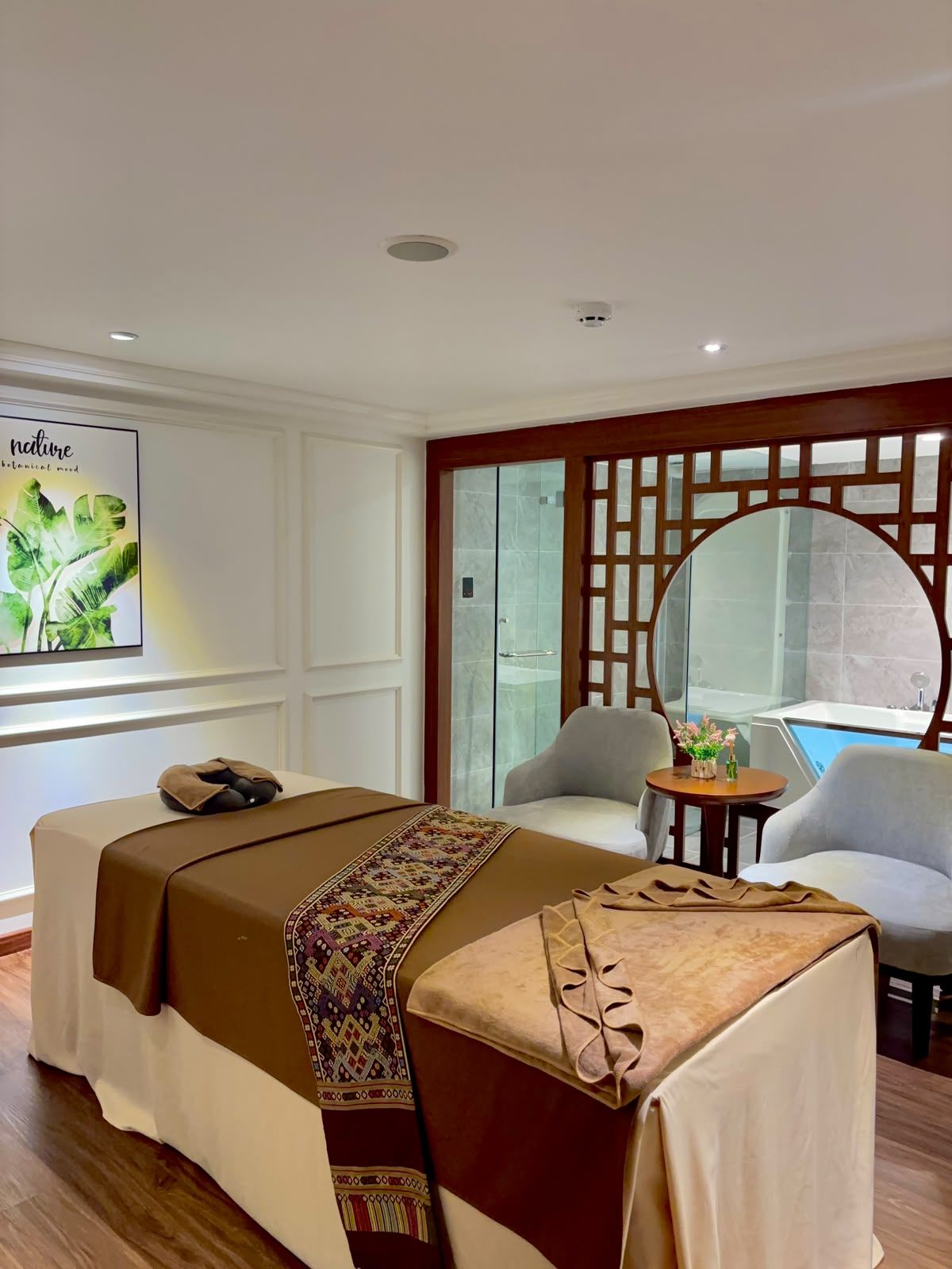 A hotel room with a massage table, two chairs, a small table with flowers, a large mirror, and a poster on the wall.